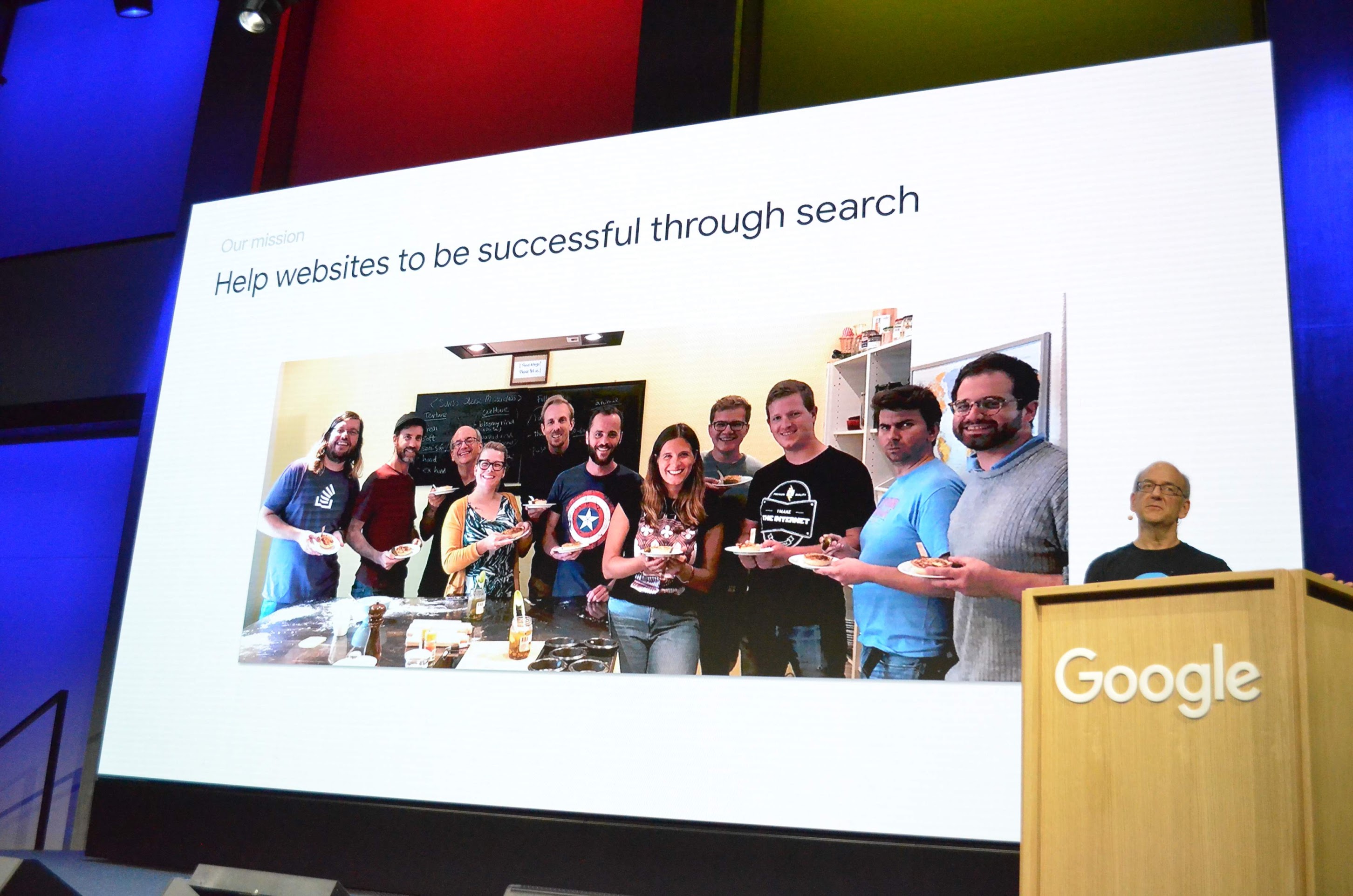 Una foto del evento Search Central Live de Zúrich en 2019 con John Mueller frente a una diapositiva con el título &quot;Help websites to be successful through Search&quot; y una foto del equipo ampliado de Relaciones de la Búsqueda de Google.